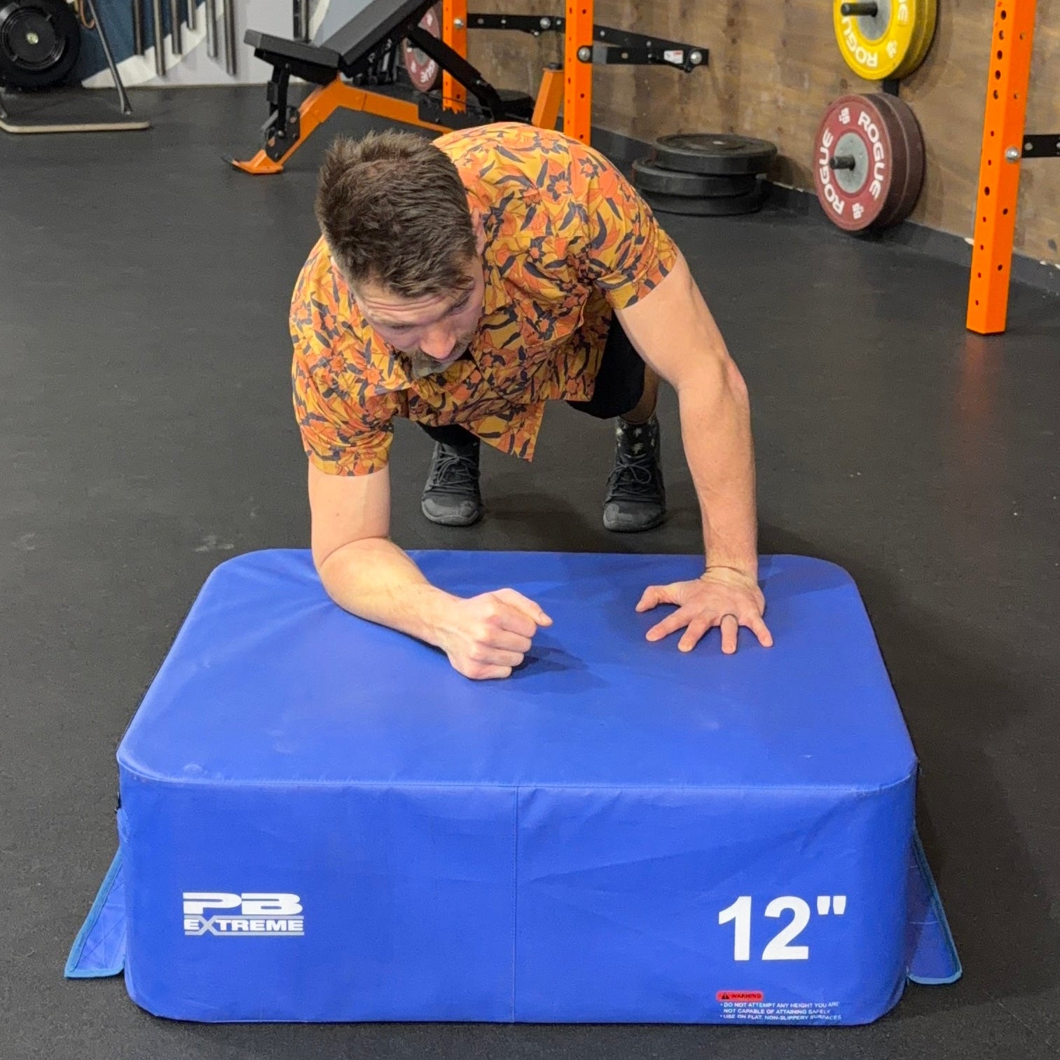 Person performing push-ups on a blue 12-inch exercise platform in a gym setting.