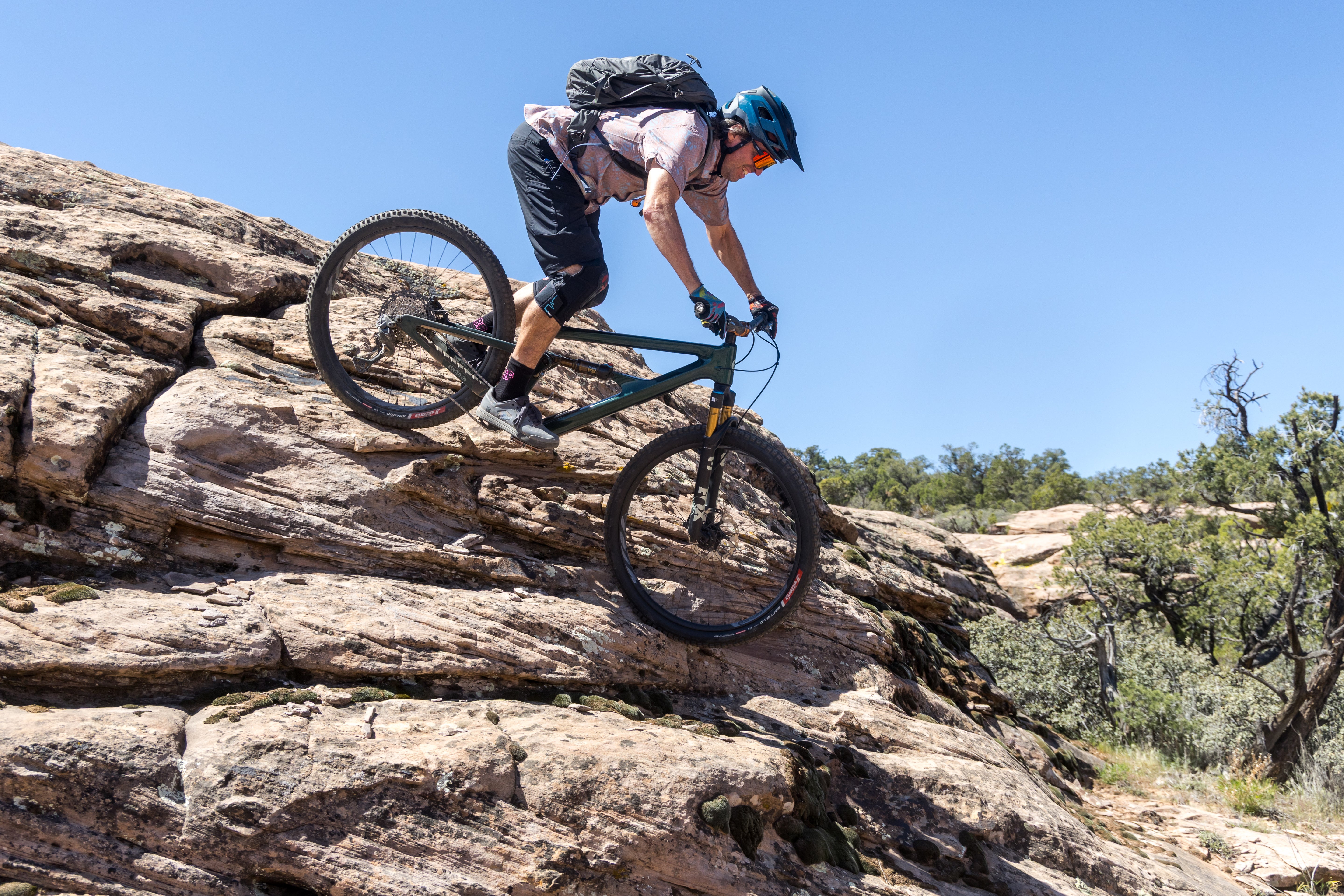 A mountain biker wearing a helmet and backpack maneuvers a black full-suspension bike over a steep rocky outcrop under a clear blue sky.
